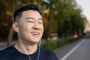 Close-up photo of a young Asian male athlete in headphones standing on a city street with his eyes closed and resting from training.