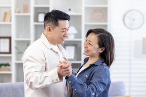 contento joven asiático familia, hombre y mujer son contento a moverse dentro un nuevo casa, abrazo, sostener manos, bailar, Mira a cada otro sonriente. foto