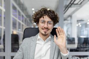 A professional young adult businessman in smart attire waves hello, showing positivity and confidence amidst a corporate setting. photo