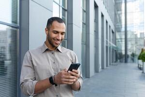 hispanic man outside modern office building using smartphone, businessman in shirt typing message and browsing online pages photo