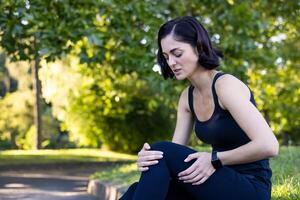 foto de un joven mujer haciendo Deportes y trotar en el parque, sentado en el bordillo y participación su pierna con su manos. siente grave rodilla dolor y esguince.