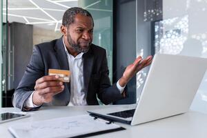 trastornado y preocupado empresario a lugar de trabajo, hombre engañado recibido en línea dinero transferir error, africano americano hombre infeliz sentado con ordenador portátil dentro oficina participación banco crédito tarjeta. foto