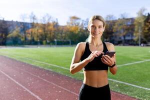 Young female sports coach standing in stadium and using phone, looking at camera smiling. photo