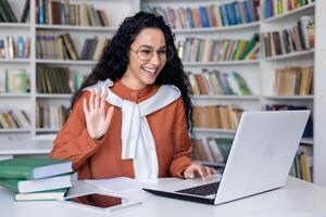 simpático contento Hispano hembra profesor conduce un en línea lección remotamente sentado a un mesa en frente de un computadora portátil, joven morena oficina trabajador es teniendo un llamar, ondulación a el cámara. foto