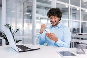 joven Hispano empresario sentado en el oficina a el escritorio y utilizando el teléfono. él mira a el pantalla, marca, se regocija, celebra, muestra un victoria gesto con su mano. foto