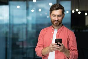 sonriente y contento empresario dentro el oficina por el ventana usos el teléfono un maduro hombre con un barba mira a el teléfono inteligente pantalla, navega Internet páginas, tipos mensajes en un en línea solicitud foto