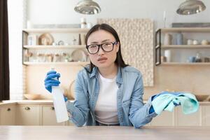 Tired young beautiful Asian woman cleaning at home. Wipes the table with a rag in rubber gloves and a bottle of spray. He looks at the camera, spreads his hands. photo