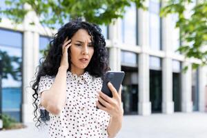 Sad disappointed woman received online notification with bad news on her phone, businesswoman walking outside office building, using application on smartphone, reading social media unsatisfied. photo