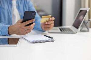 de cerca de un del hombre manos utilizando un teléfono inteligente y crédito tarjeta a hacer un en línea compra con ordenador portátil y cuaderno en antecedentes. foto