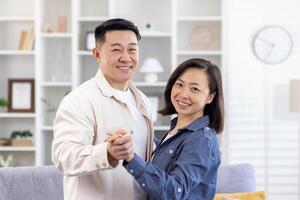 Portrait of young happy Asian couple hugging, holding hands at home dancing, man and woman looking at camera smiling. photo