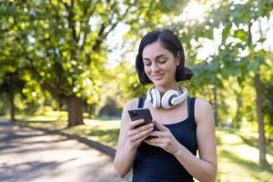A cheerful young lady wearing headphones around her neck, using a smartphone in a leafy park setting. photo