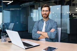 Portrait of mature businessman with beard, man in shirt smiling and looking at camera sitting at desk with crossed arms inside modern office building, using tablet and laptop at work. photo