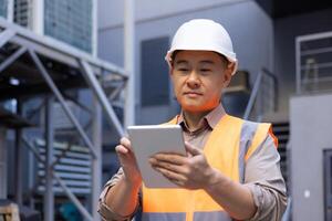 Focused Asian engineer with hard hat using digital tablet at industrial site. Technology and engineering concept. photo