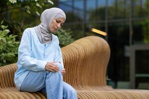 Muslim female sitting outside and raising leg bent in knee with suffering expression on background of modern building. Sick woman facing trauma consequences and rubbing injured place with both hands. photo