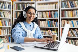 sonriente mujer en un biblioteca ajuste utilizando un ordenador portátil con auriculares. ella es rodeado por expansivo estantería, ejemplificando un productivo investigación ambiente. foto