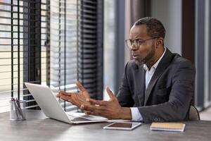 Clueless business person spreading hands to computer while working by table with tablet and notebook. Mature man in jacket trying solving company processes and feeling desperate about new tasks. photo