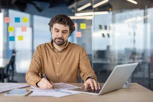 enfocado empresario analizando documentos mientras utilizando un ordenador portátil a un escritorio en un contemporáneo oficina ajuste con pegajoso notas en el antecedentes. foto