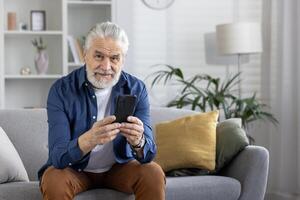 alegre mayor hombre con un barba sonriente mientras utilizando un teléfono inteligente cómodamente sentado en un sofá a hogar. foto