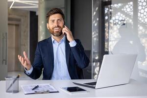 sonriente joven negocio hombre teniendo un negocio conversacion en móvil teléfono con un cliente, sentado en el oficina en un traje a el mesa y gesticulando con su manos. foto