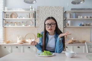 Portrait of young dissatisfied Asian woman eating salad at home and looking at camera. photo