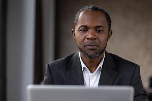 Portrait of concerned diverse man looking attentively into monitor of computer on blurred background. Strict employer in shirt checking productivity of marketing department remotely from cabinet. photo