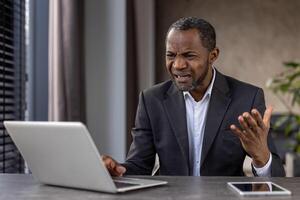 Annoyed black male looking at laptop screen with confusion and raising hand in despair while sitting at home office. Angry businessman can not continuing working due to technical problems. photo