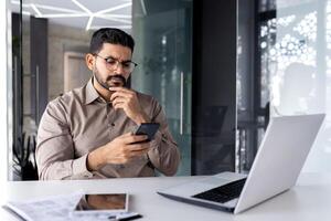 empresario pensando mientras sentado dentro oficina a mesa, hombre es concentrando y seriamente leyendo en línea utilizando un solicitud en teléfono, el joven jefe es participación un teléfono inteligente en su manos. foto