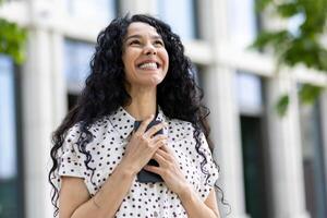 optimista joven latín americano mujer con Rizado pelo sonriente fuera de un oficina edificio, reflejando confianza y éxito. foto