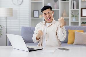 Asian working remotely from home office with laptop, businessman received notification of good achievement results, freelancer satisfied with work rejoices and celebrates victory and triumph. photo