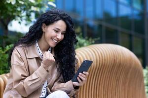 Young happy woman celebrating victory triumph success, satisfied hispanic business woman holding phone received online win notification, sitting on bench outside office building. photo