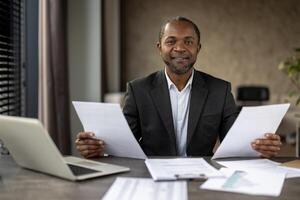 Calm african american consultant organising paperwork while sitting at comfortable workplace in neutral colours. Relaxed professional checking accuracy and reliability of contract for client. photo