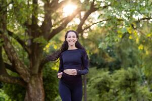 Young beautiful woman running in the park, portrait of a Latin American woman jogging and doing active exercises, sportswoman smiling and looking at the camera among the trees. photo