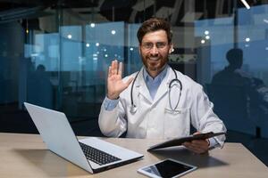 retrato de exitoso maduro médico dentro moderno clínica, hombre trabajando con ordenador portátil mirando a cámara y sonriente ondulación mano saludo gesto foto