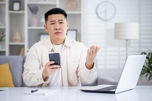 Disturbed chinese male in casual clothes holding mobile phone while working at home office with portable computer. Unhappy remote manager receiving text message with penalty in corporate chat. photo