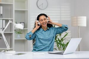 A delighted woman in a blue shirt listens to music with headphones, smiling broadly as she enjoys a break at her clean, contemporary office workspace. photo