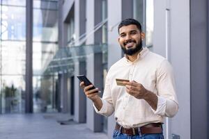 A casually dressed young adult is engaged with his smartphone while holding a credit card on a busy urban street, suggesting online shopping or financial management. photo