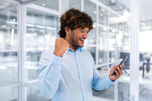 Successful hispanic programmer inside office and celebrating victory and successful achievement, man reading received news online phone, businessman in shirt holding hand up triumph gesture photo