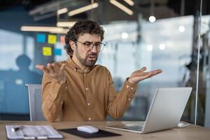 un adulto masculino en un casual camisa expresa Confusión mientras utilizando un ordenador portátil en un contemporáneo oficina configuración, reflejando común lugar de trabajo retos foto