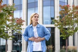 maduro mujer en un camisa es caminando en el ciudad, un trabajador tiene un grave estómago dolor, un rubia mujer de negocios en casual ropa es enfermo, masajear su manos con su manos fuera de un oficina edificio. foto