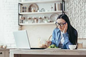 Anorexia. Dissatisfied and tired young Asian woman trying to diet. Sitting in the kitchen with a laptop and eating fresh salad, vegetables. She feels bad, holds her head with her hand, grimaced. photo