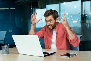 Angry and nervous businessman talking on call remotely, man shouting to colleagues online, boss in shirt working inside modern office using laptop at work. photo