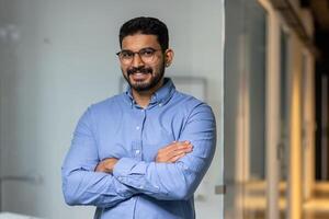 retrato de un alegre masculino profesional en un azul camisa, cruce brazos con confianza en un moderno lugar de trabajo. foto