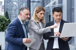 Successful business team, three colleagues businessman and businesswoman outside office building discussing current plans and management, looking at laptop screen, discussing and consulting photo