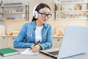 Online training. Young beautiful Asian girl sitting at the table at home in white headphones and with a laptop. He studies remotely. Writes down a lecture, class in a notebook. photo