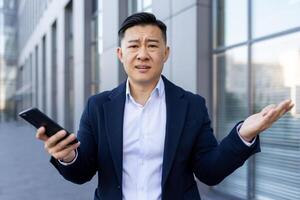 Close-up portrait of a young Asian male businessman standing near an office building and upset, spreading his hands, holding the phone and looking worriedly at the camera. photo