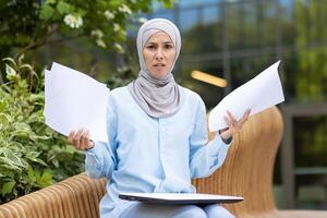 Muslim businesswoman with hijab holding papers with a puzzled expression, working outdoors on a warm day. photo