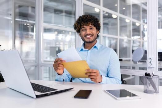 Portrait of a happy Hindu programmer inside the office, a businessman holds an envelope with a good news message in his hands, the man smiles and looks at the camera, uses a laptop at work. photo