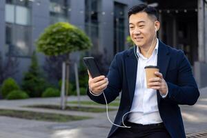 Asian young male office worker sitting outside an office building wearing headphones, talking on the phone on a call and drinking coffee. photo