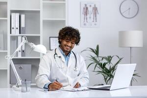 joven médico trabajos con un ordenador portátil dentro el médico oficina de el clínica, el hombre felizmente sonrisas y llena fuera médico informes y tratamiento formularios, trabajos con documentos. foto