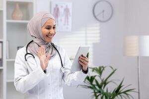 un alegre musulmán cuidado de la salud profesional en un hijab se involucra con pacientes virtualmente en un moderno clínica ajuste. foto